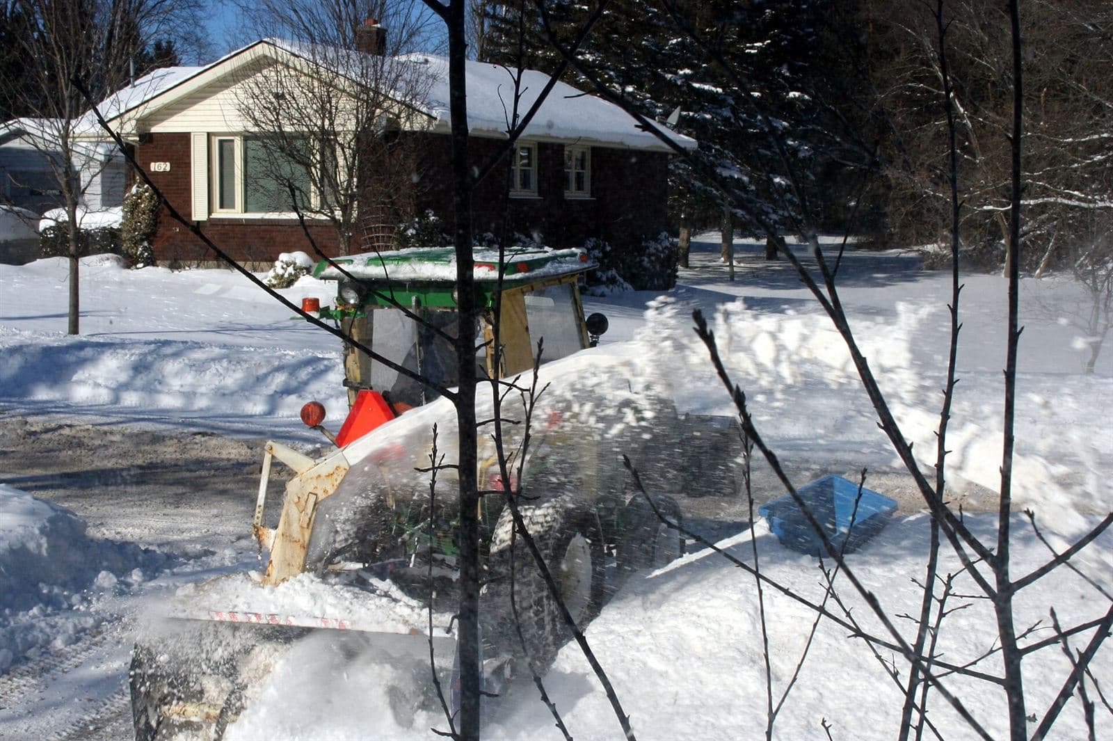 Tractor snowblower clearing deep snow from a residential driveway beside a brick bungalow in Ontario
